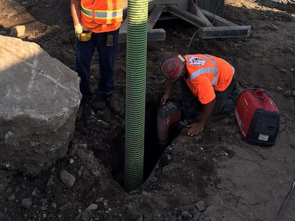 Vacuum hose lowered into a narrow excavation while a worker loosens soil at the bottom
